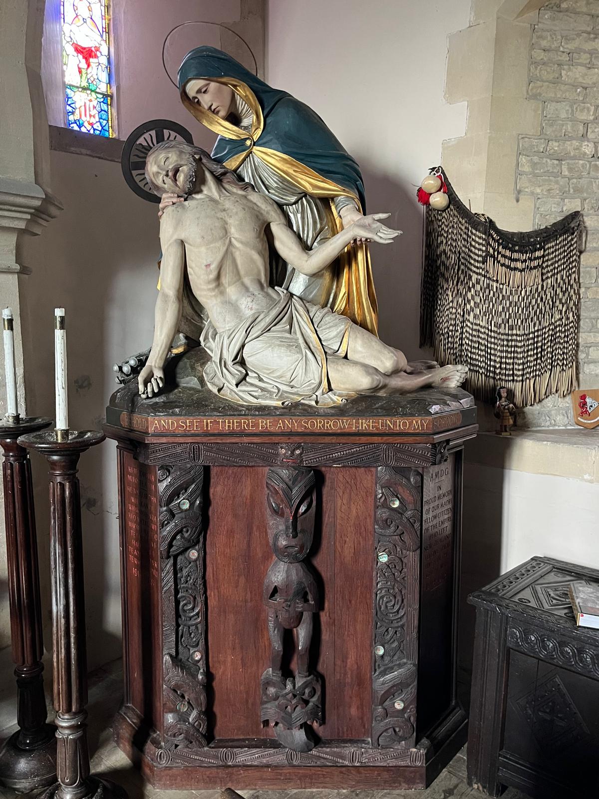 A wooden memorial with a painted figure of Mary holding the body of Christ mounted on a plinth with supports made up of carved Māori figures.