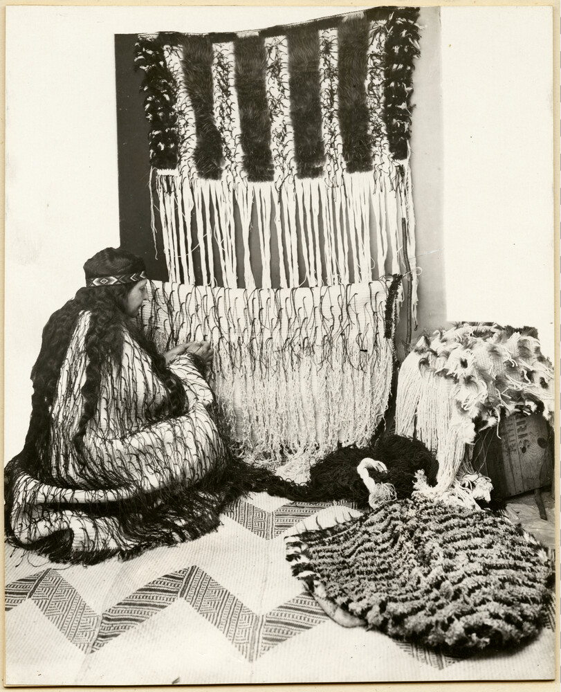 Makareti crouches on a mat and weaves a korowai cloak next to a whariki with a rahokuia resting on top.