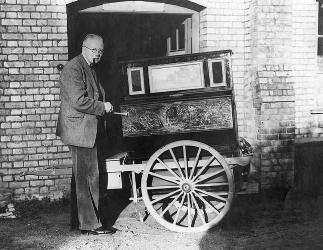 A black and white photo of TK Penniman standing in a suit and holding the handle of a portable street piano.