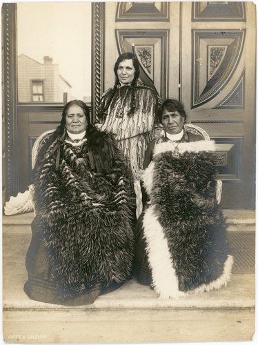Three Maori women in feather cloaks are posed in front of a backdrop of panelling.